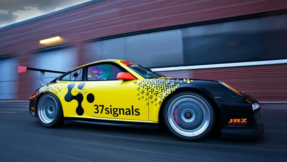 A yellow and black Porsche race car with 37signals branding and a rear wing speeding past a brick building.