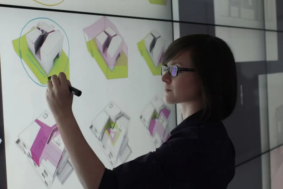 A person in glasses draws a circle with a stylus on a large wall-mounted touchscreen displaying product sketches.