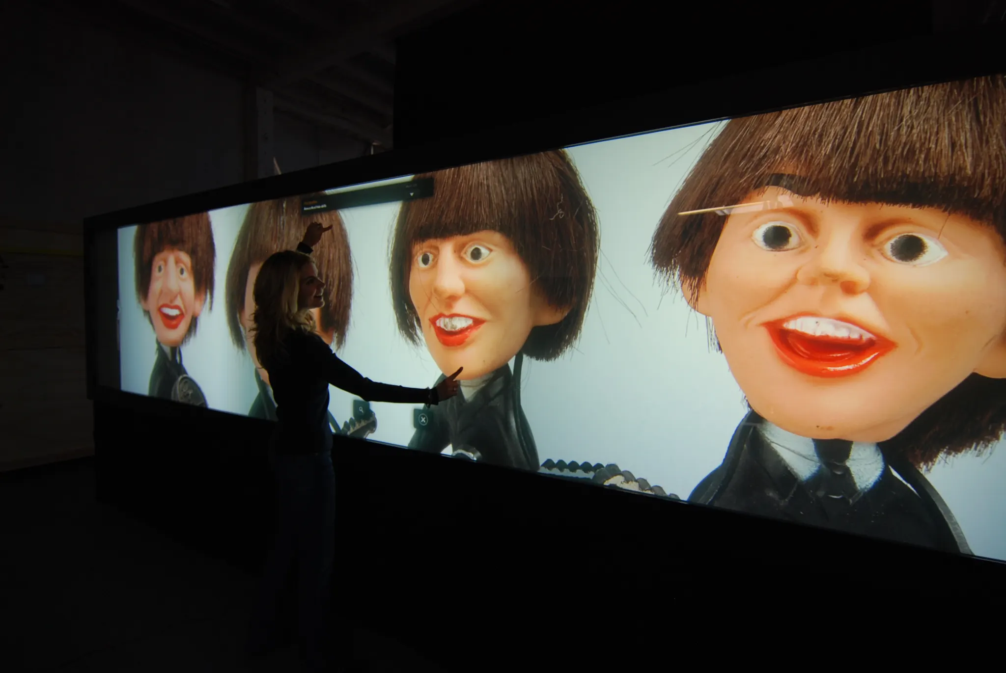A person stands touching a long dark projection wall showing large mop-top bobblehead doll faces in a dim room.