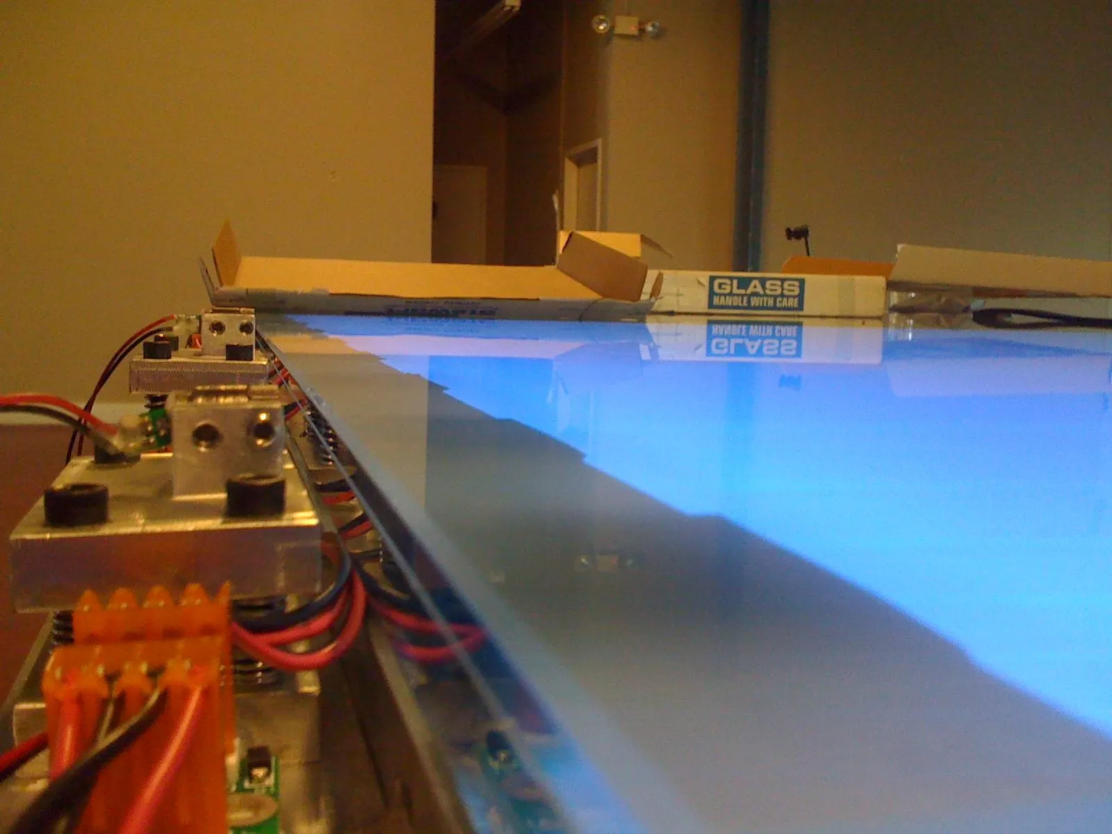 A large glossy table surface glowing blue at close angle, with exposed circuit boards, wiring, and metal mounts along its edge.