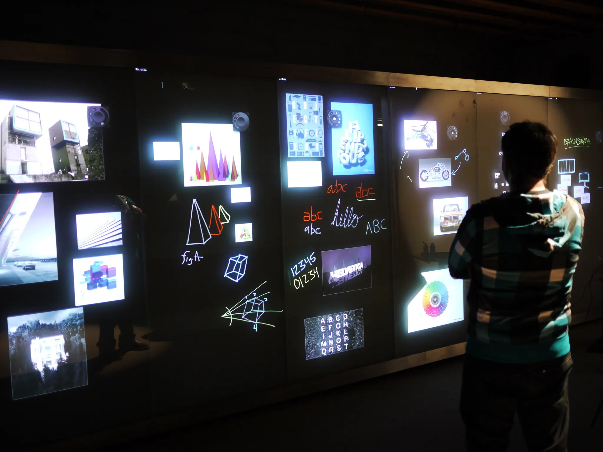 A person stands facing a large dark interactive wall covered in glowing images, sketches, and handwritten text.