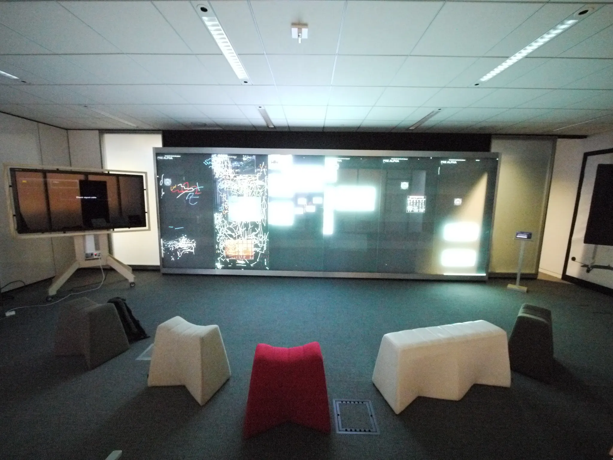 A dim room with a large multi-panel wall display showing a bright interface, faced by white and red angular seating.
