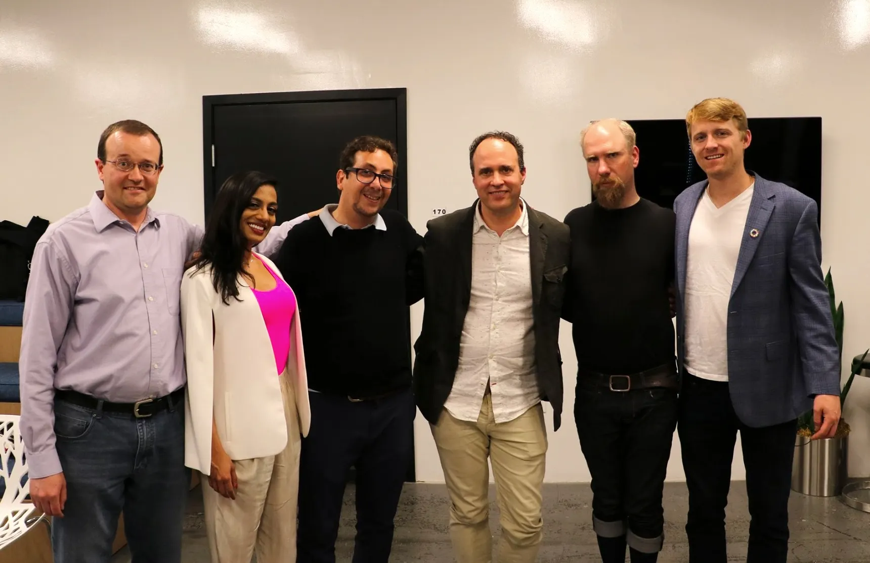 Six people stand together in a row indoors, arms around each other, in front of a white wall and dark doors.