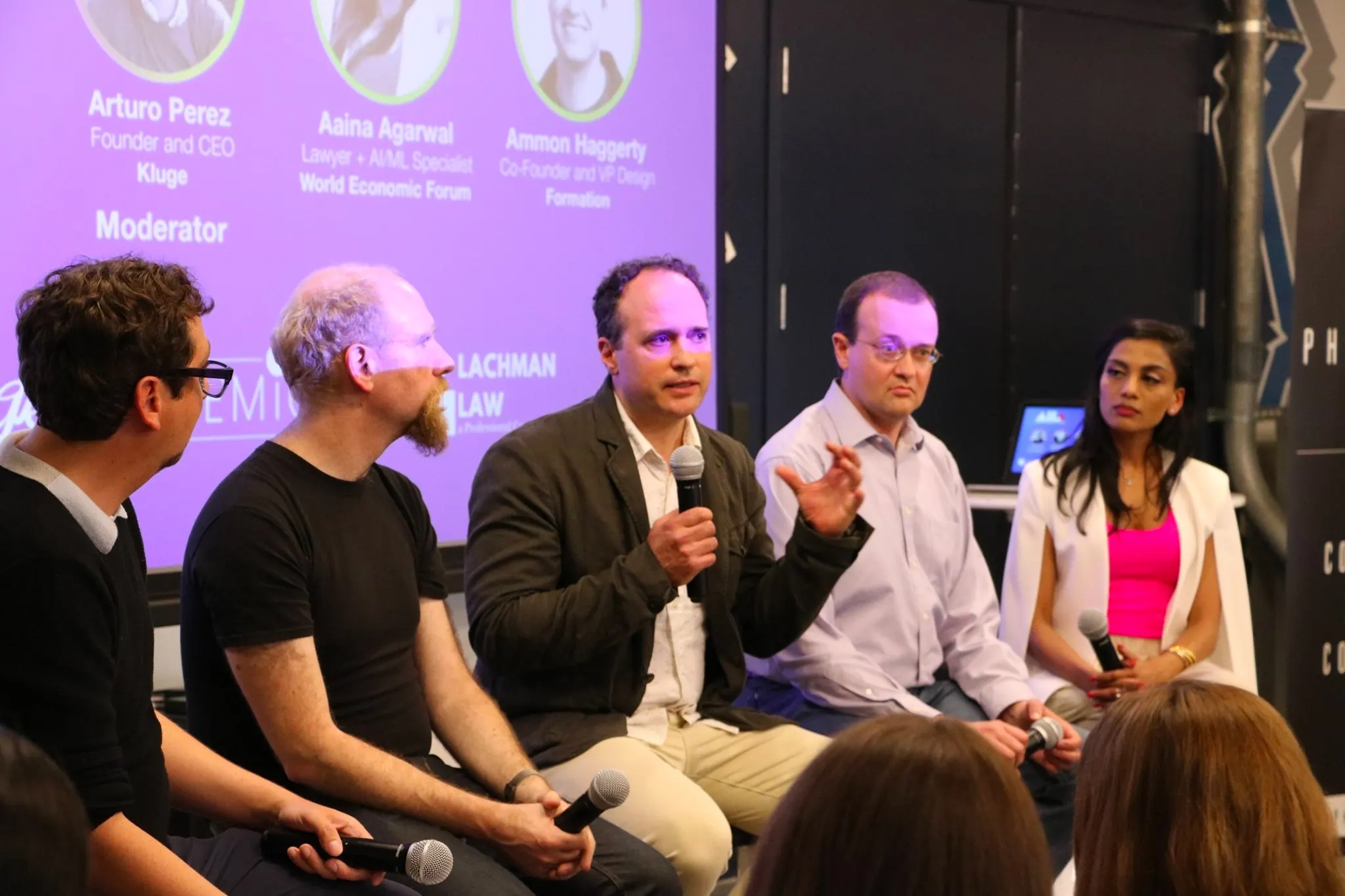 Five panelists seated in front of a purple projection screen listing speaker names, one holding a microphone and speaking.