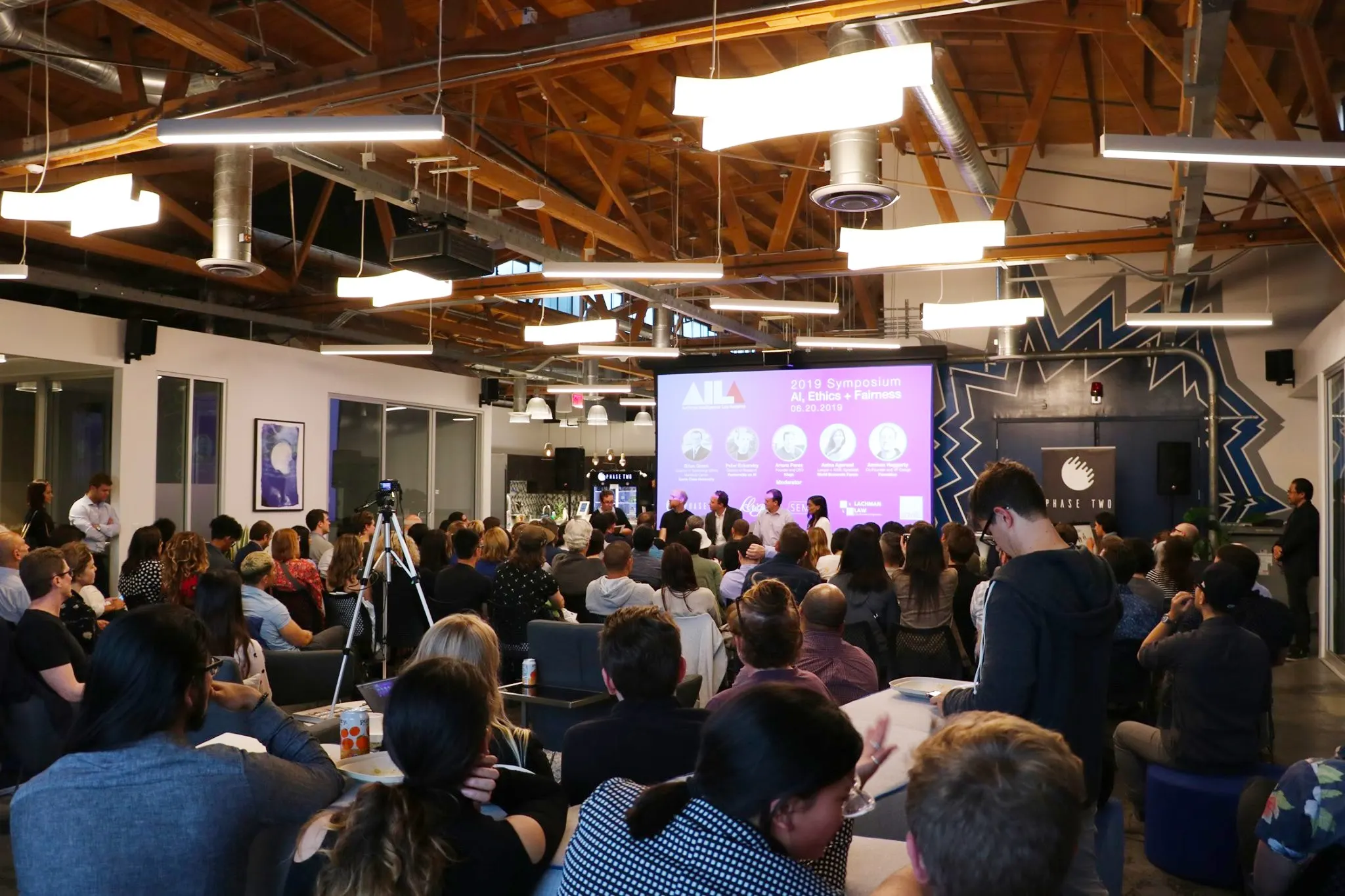 A seated audience in an open loft space with wood beams watches a panel on stage beside a large projection screen.