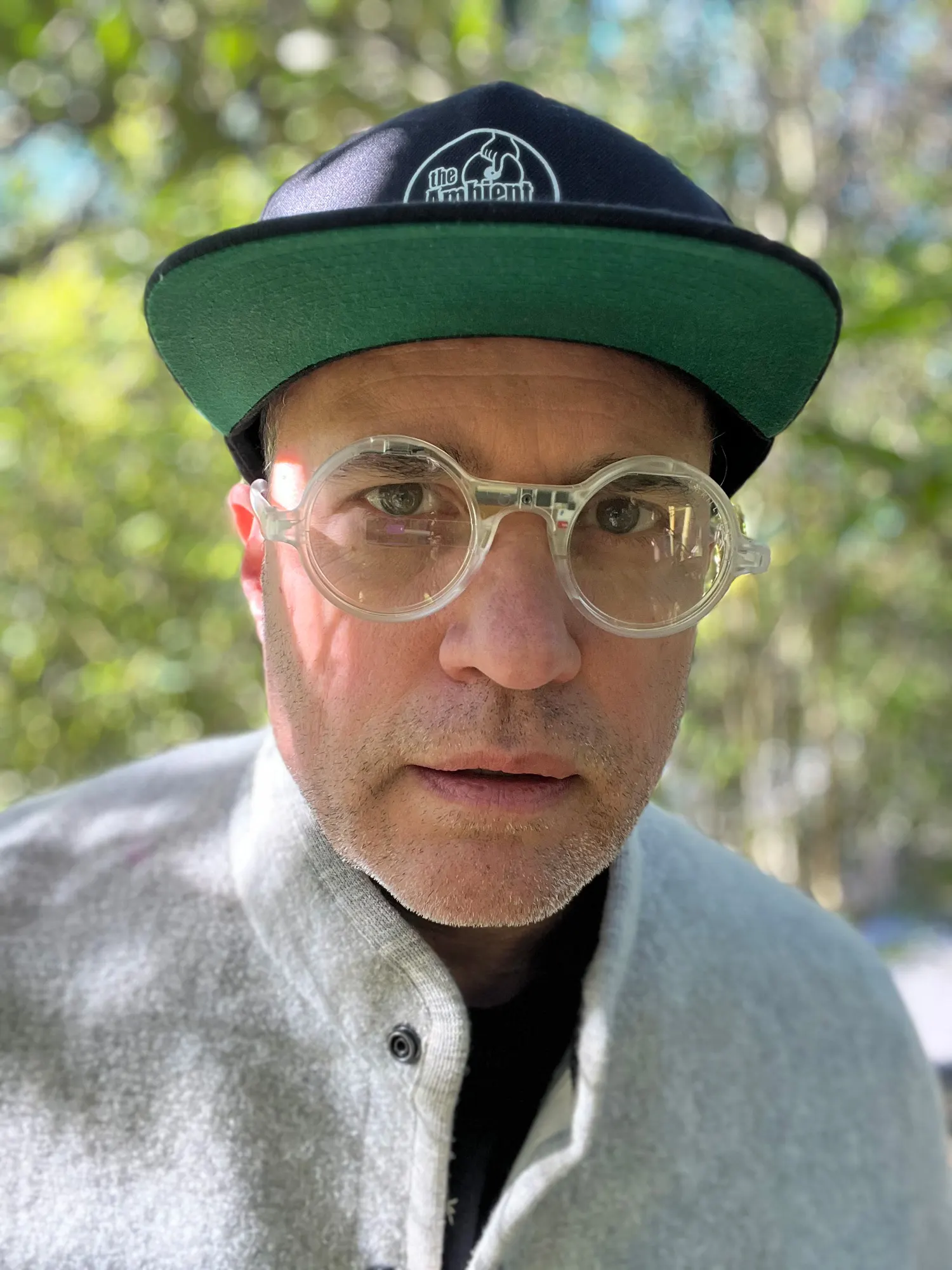 Close-up portrait of a person in clear round glasses, a green and navy cap, and a grey fleece jacket outdoors.
