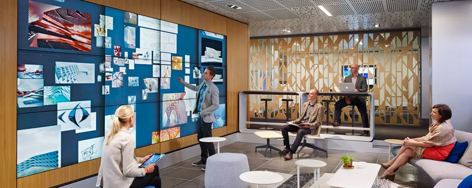 A person presents on a large multi-screen video wall of images in a modern office lounge as three others watch from seats.