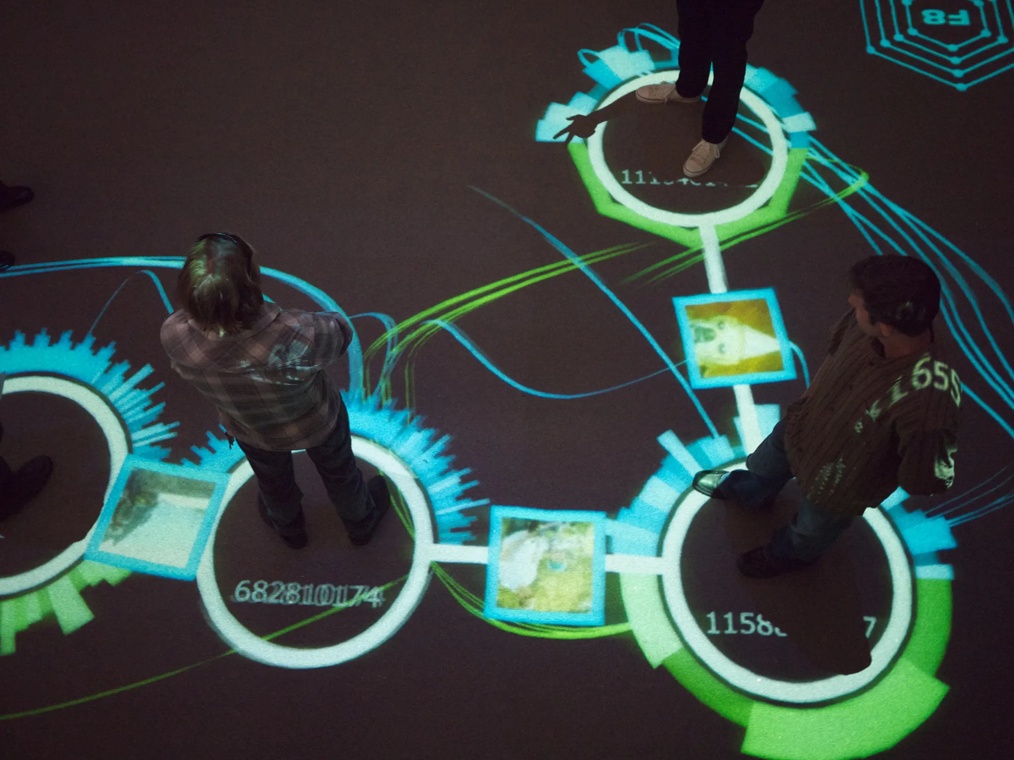 Overhead view of people standing on a floor projection of glowing cyan and green rings, curved lines, numbers and photos.