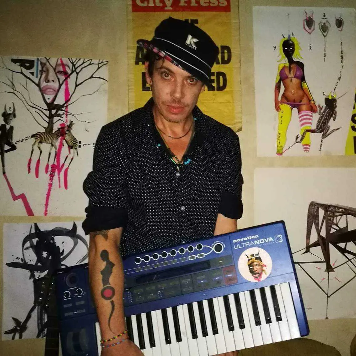 A person in a bucket hat and polka-dot shirt holds a blue Novation UltraNova synthesizer in front of artwork-covered walls.