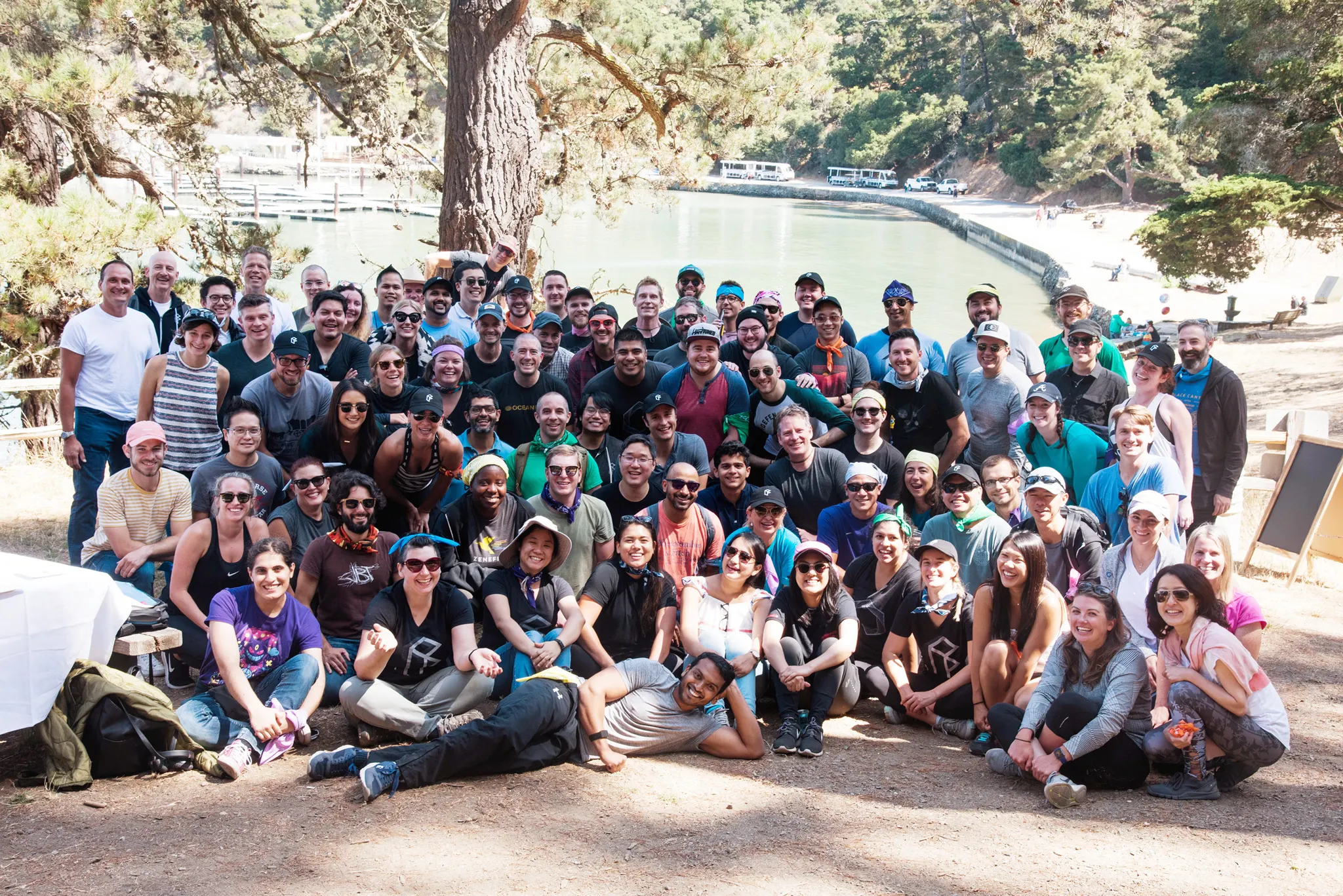 A large group photo of dozens of people posing outdoors on a dirt path with trees and a cove behind them.