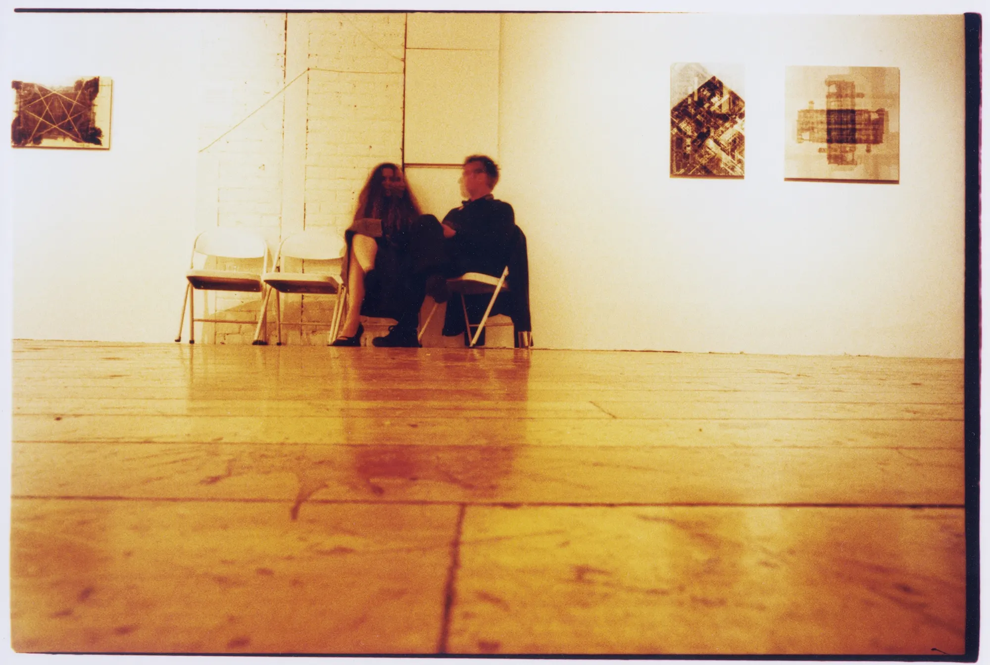 Two people sit on folding chairs in a warm-toned gallery with a wooden floor and framed abstract works on white walls.