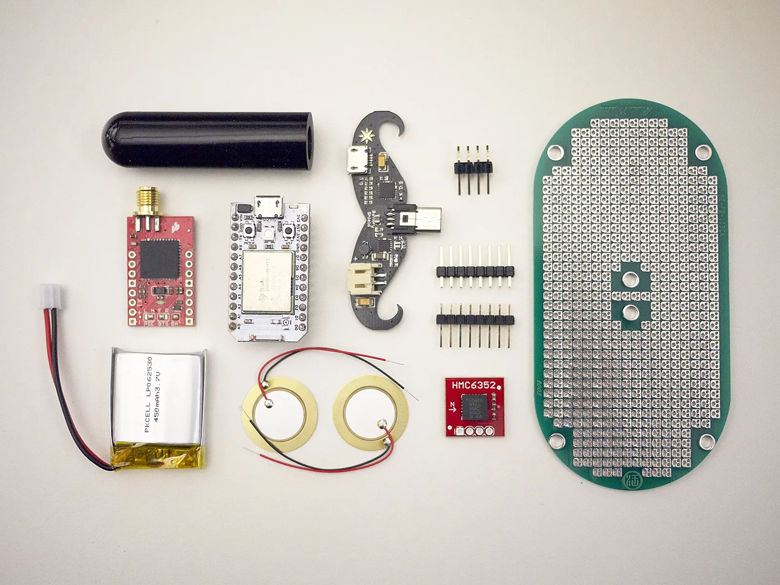 Electronic components arranged in a grid: black tube, red and green breakout boards, lithium battery, header pins, piezo discs, and a large oval perfboard.