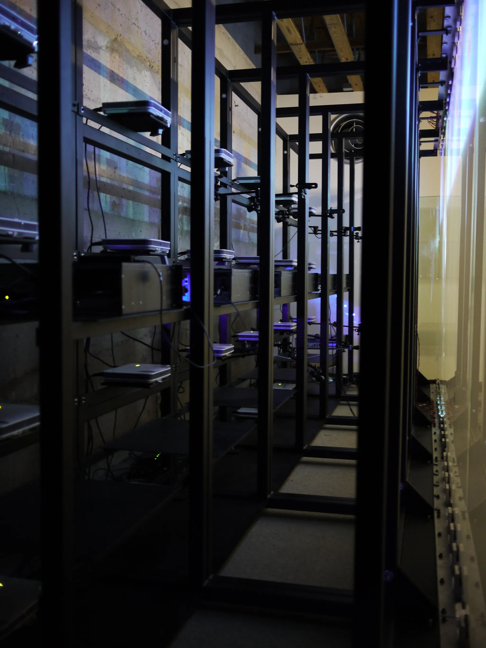 Dim narrow space behind a wall of screens, with black metal racks holding projectors and cabling.