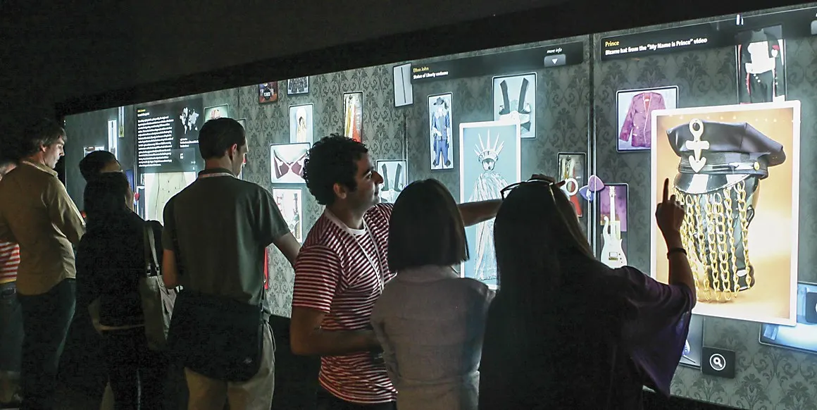 Visitors in a dim gallery gather at a large illuminated interactive wall displaying photos of costumes and memorabilia, one touching the screen.