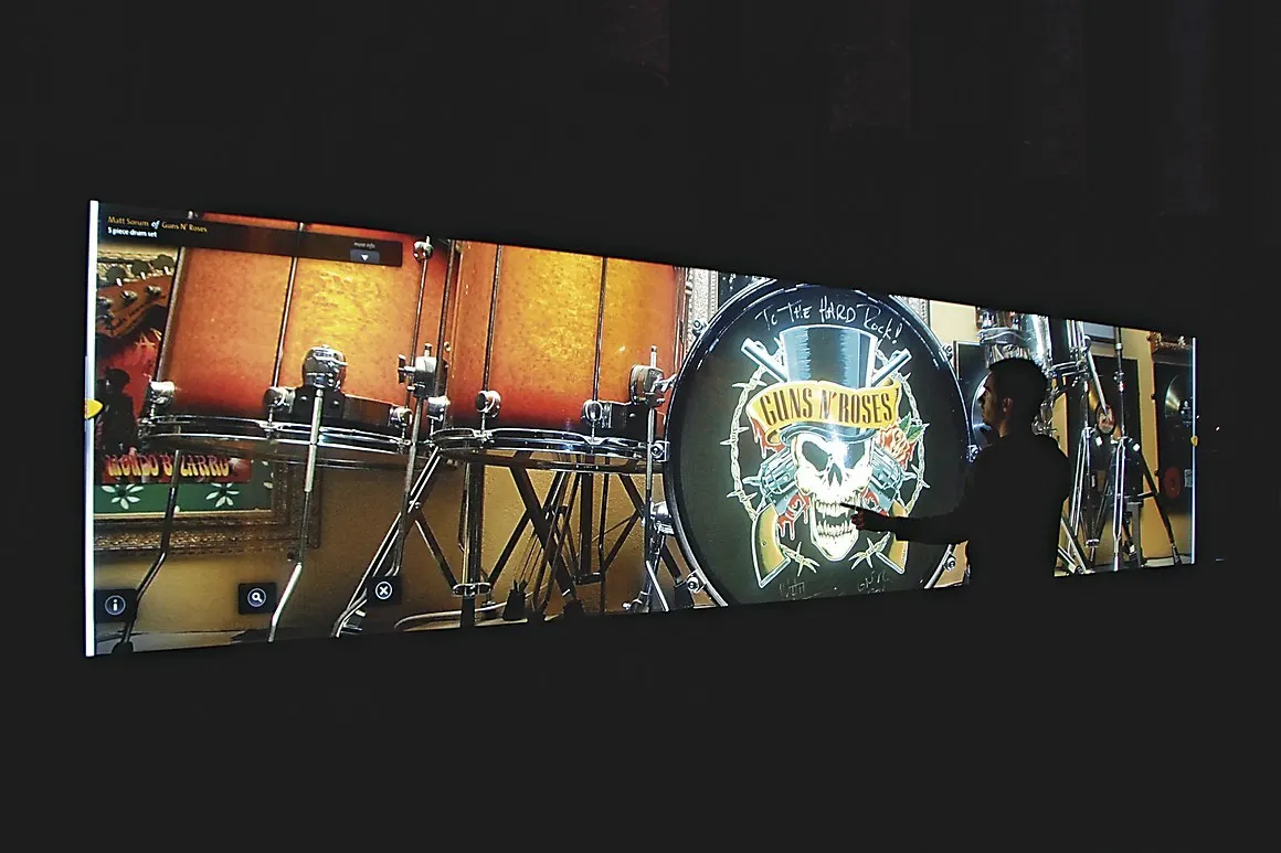 Wide panoramic wall display in a dark room showing a drum kit with a Guns N' Roses bass drum head, with a silhouetted figure in front.