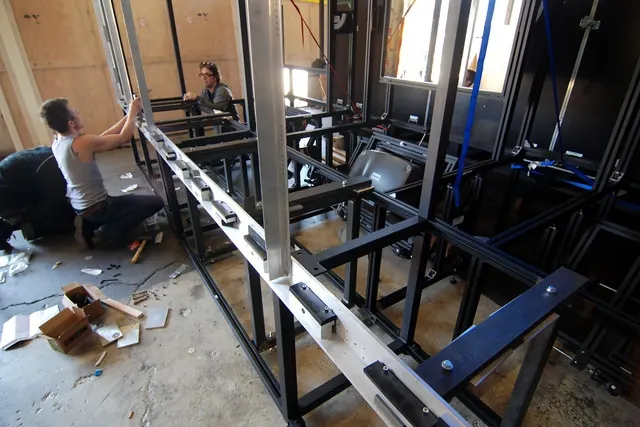 Two people assemble a large black metal frame with aluminum rails and monitors in a workshop space.