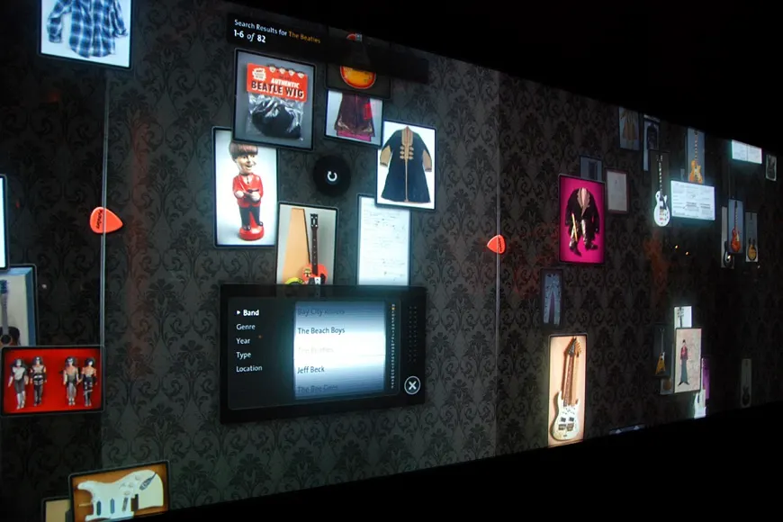 A large dark interactive video wall displaying rows of memorabilia images, guitars, and costumes with an open detail panel.