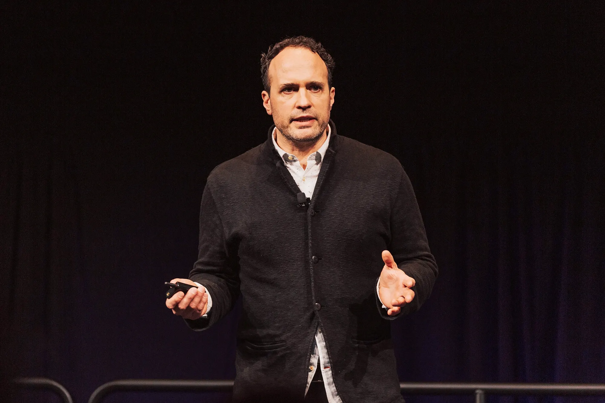 A speaker in a dark cardigan gestures on stage, holding a presentation remote against a dark backdrop.
