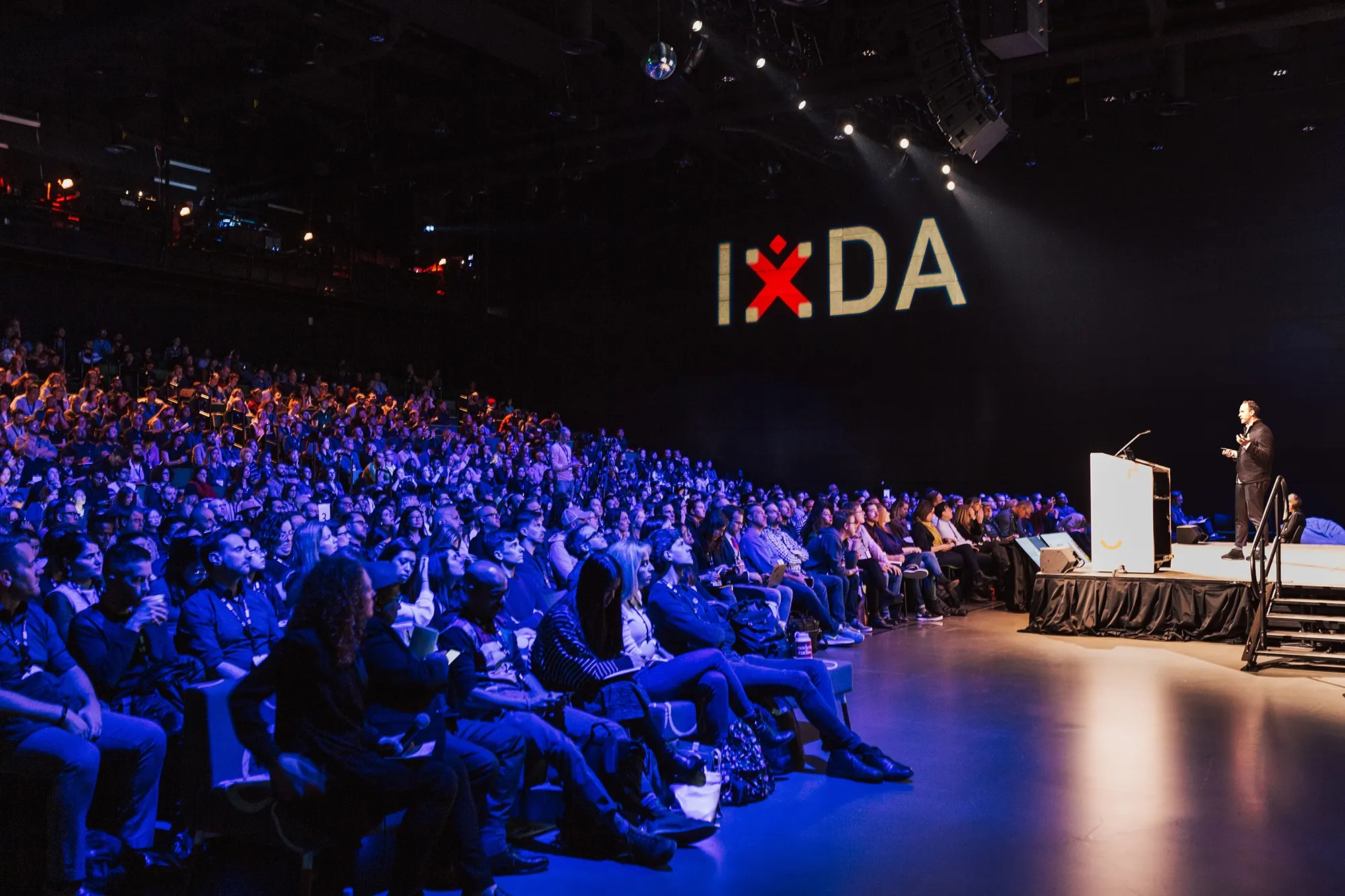 A speaker at a lectern on a dark stage addresses a large seated audience beneath a glowing IxDA sign.