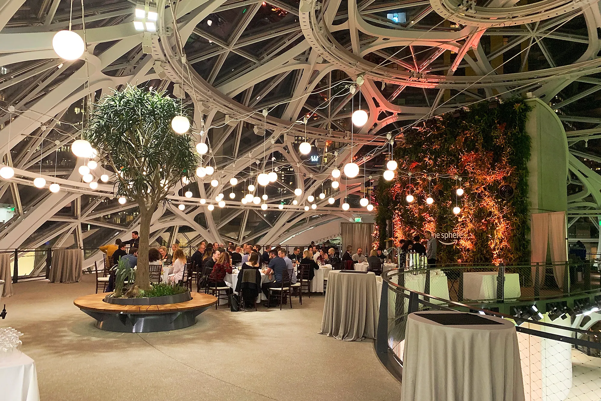 Interior of a glass-and-steel domed atrium with hanging globe lights, a tree, a green living wall, and people seated at tables.