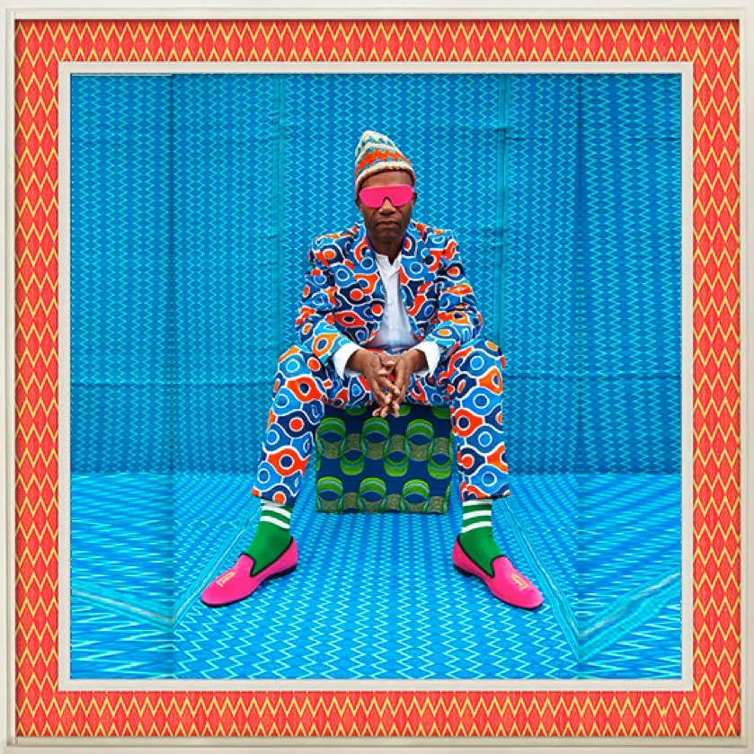 A studio portrait of a seated man in a patterned suit, knit cap, pink sunglasses and pink shoes against blue patterned cloth, in a zigzag border.