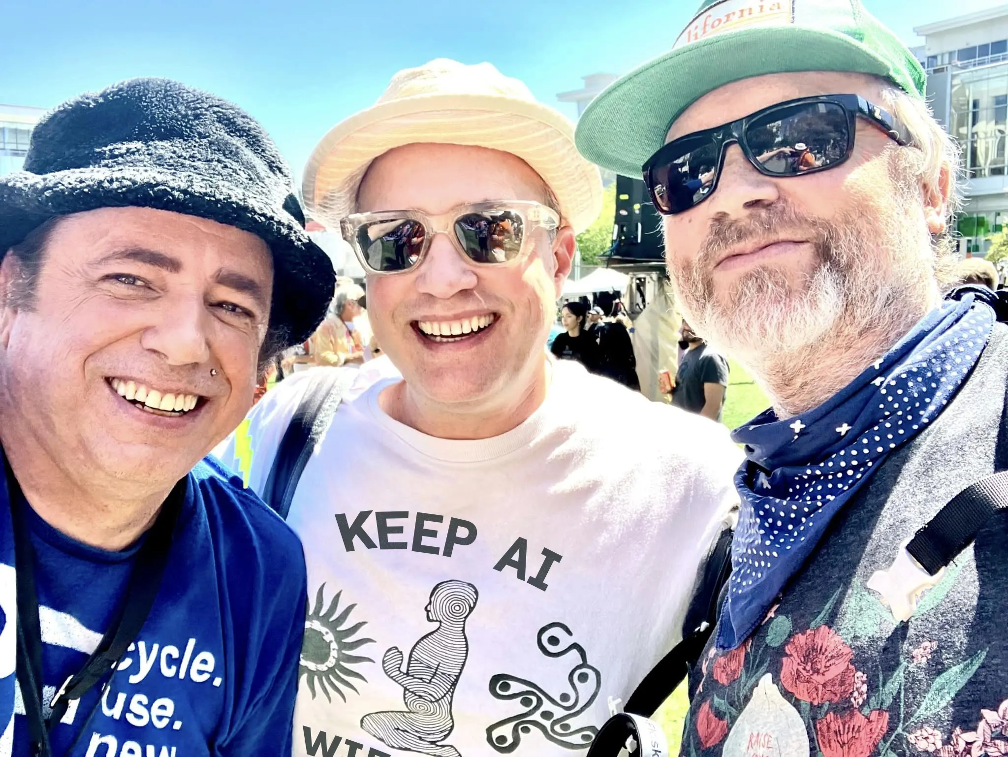 Three men in hats and sunglasses pose close together outdoors, the middle one wearing a "Keep AI Weird" t-shirt.