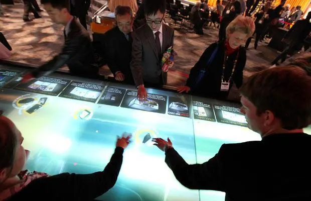 Several people stand around a large glowing multitouch table, touching cards and images on its illuminated surface.