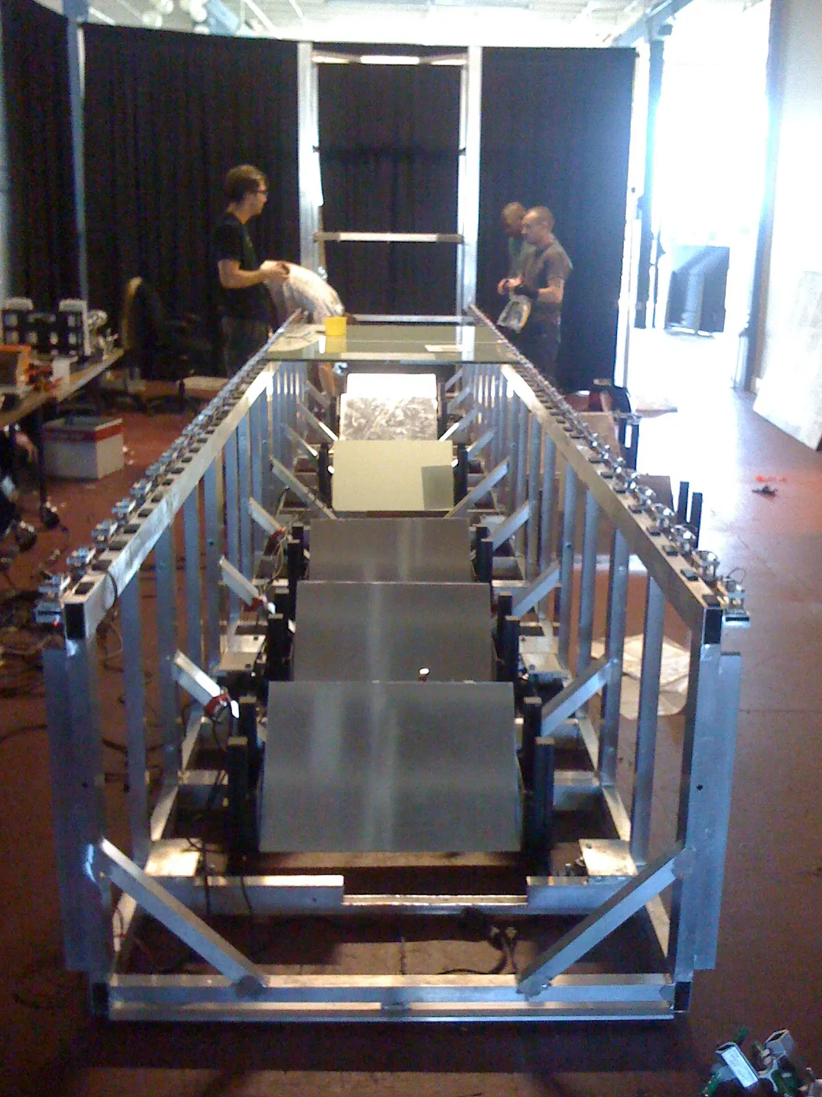 A long aluminum frame structure holding metal panels and motors under construction in a workshop, with three people working behind it.