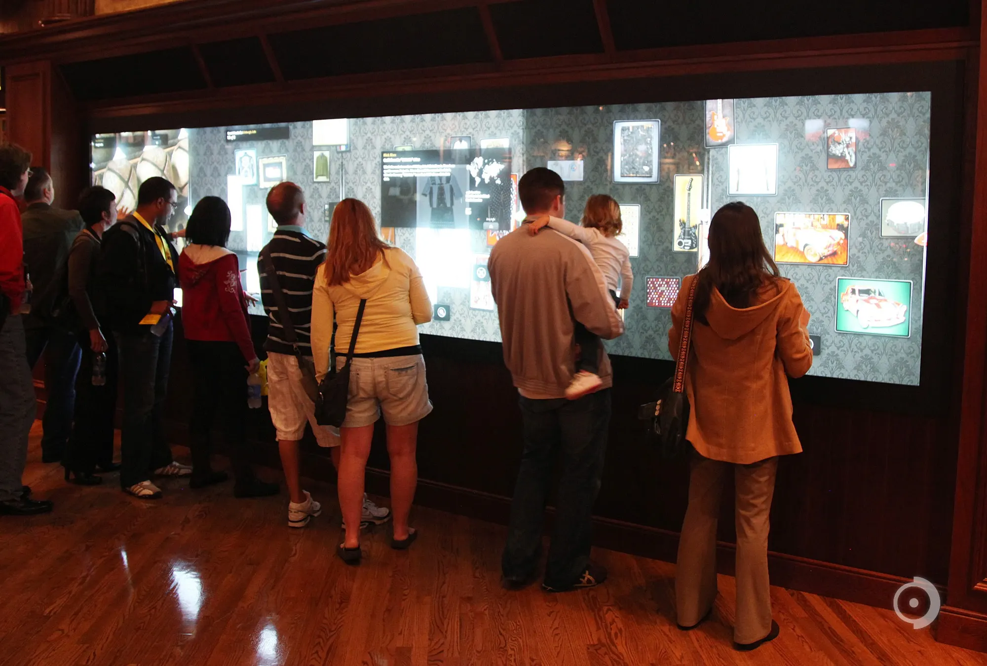 Visitors gathered along a large wall-mounted interactive touchscreen display showing photos and memorabilia in a wood-paneled room.