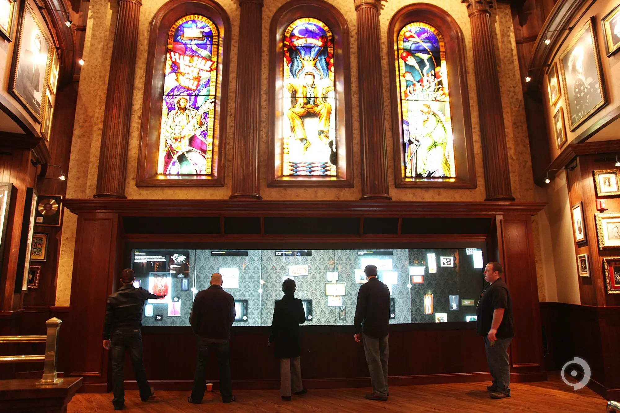 Five people stand at a long illuminated interactive display case beneath three stained glass windows in a wood-paneled room.