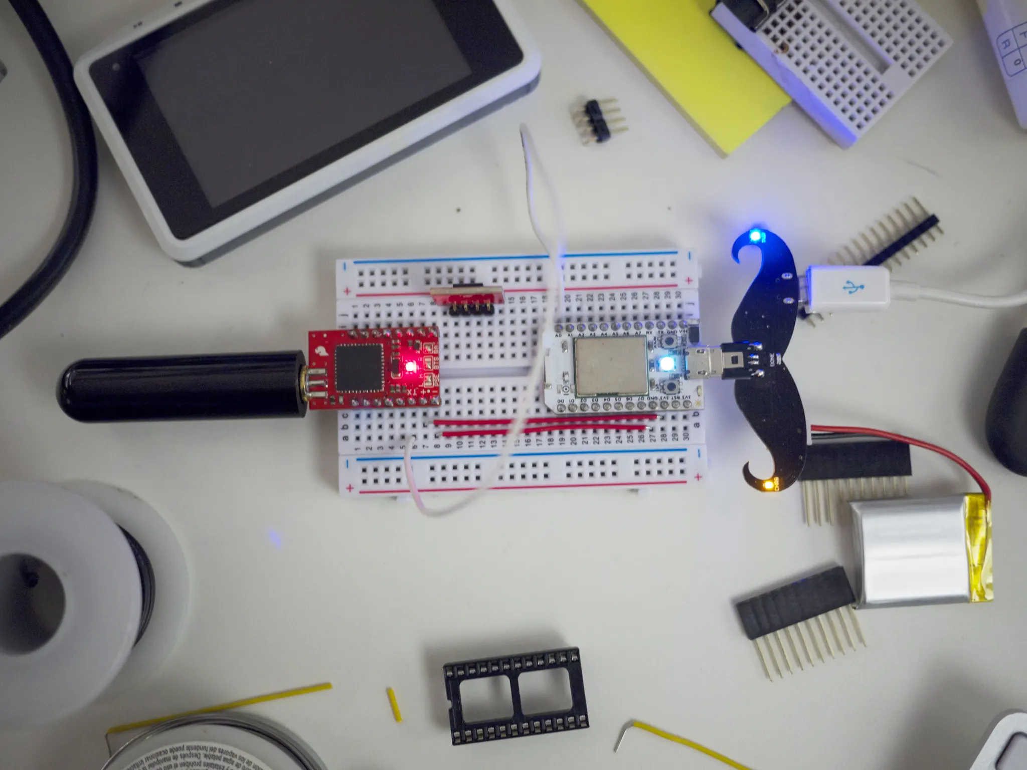 Overhead view of a white workbench with a breadboard, red microcontroller, wires, LiPo battery, header pins and a tablet.