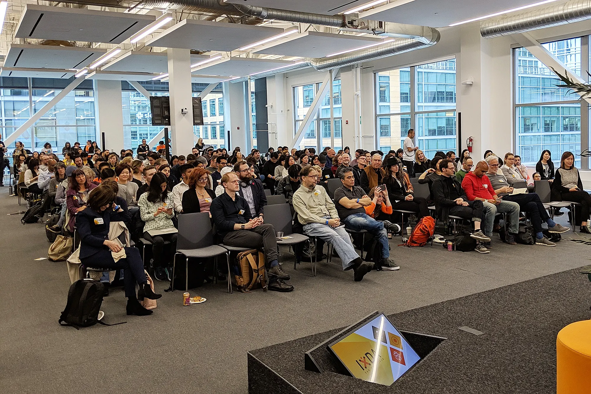 A seated audience filling rows of chairs in a bright office-like event space with large windows, viewed from the stage.