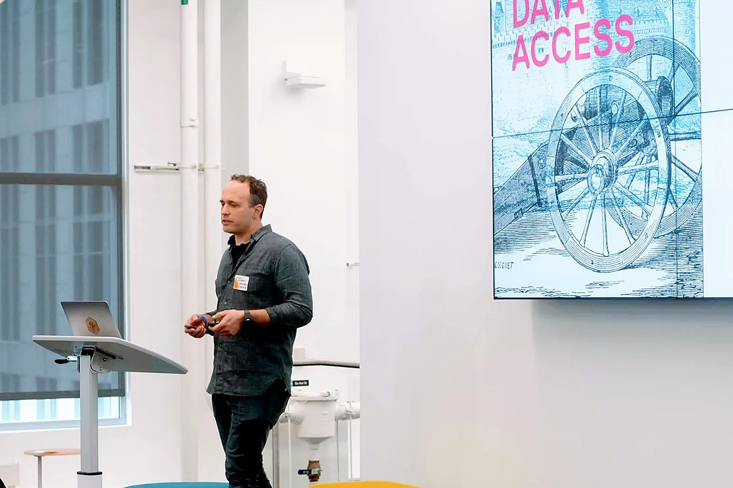 A speaker in a dark shirt stands beside a laptop on a stand, presenting near a screen reading "DATA ACCESS" over a wagon-wheel illustration.
