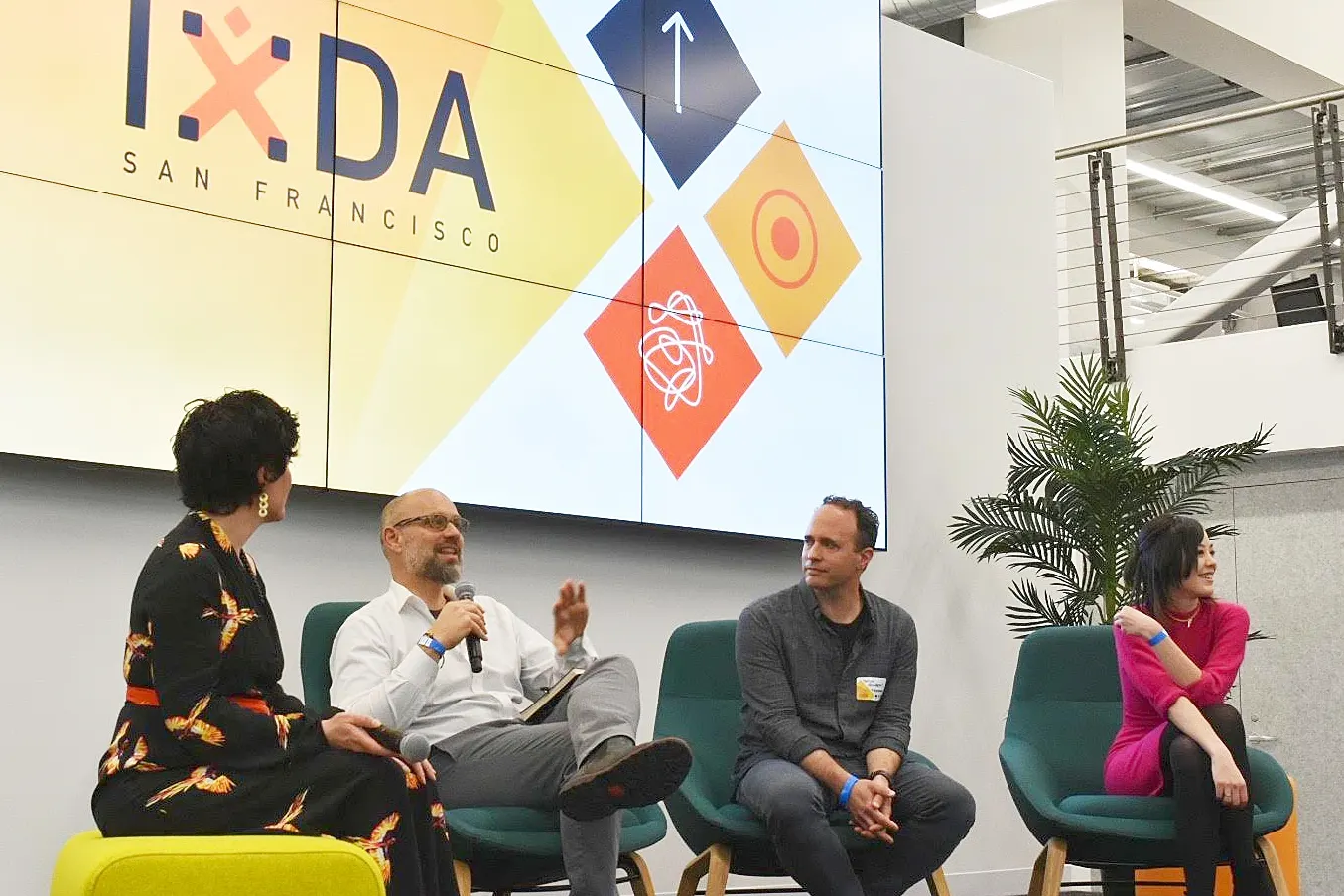 Four panelists seated in armchairs on a stage in front of a large IxDA San Francisco screen, one speaking into a microphone.