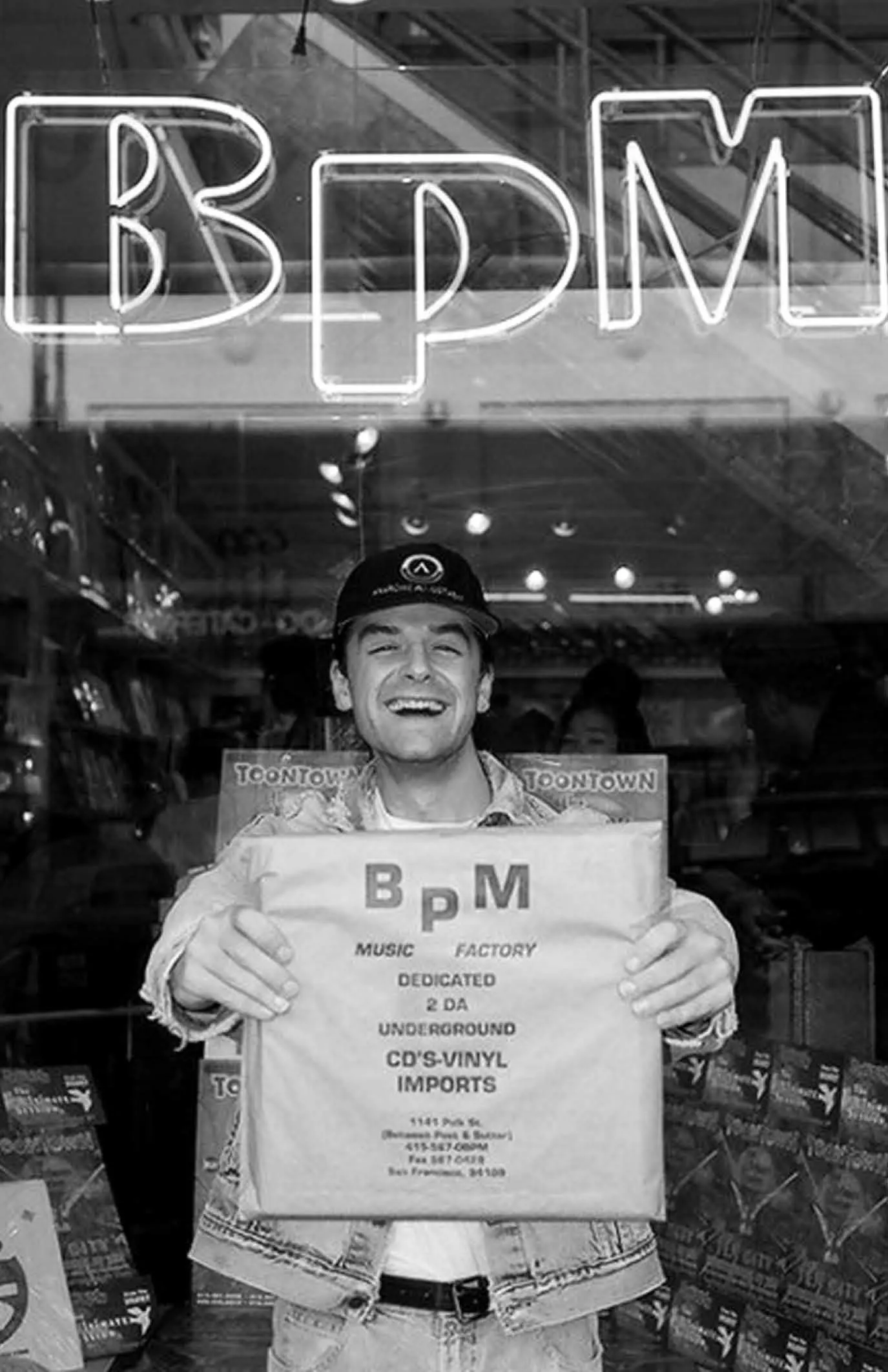 Black-and-white photo of a smiling person in a cap holding a BPM Music Factory record bag behind a window with a BPM neon sign.