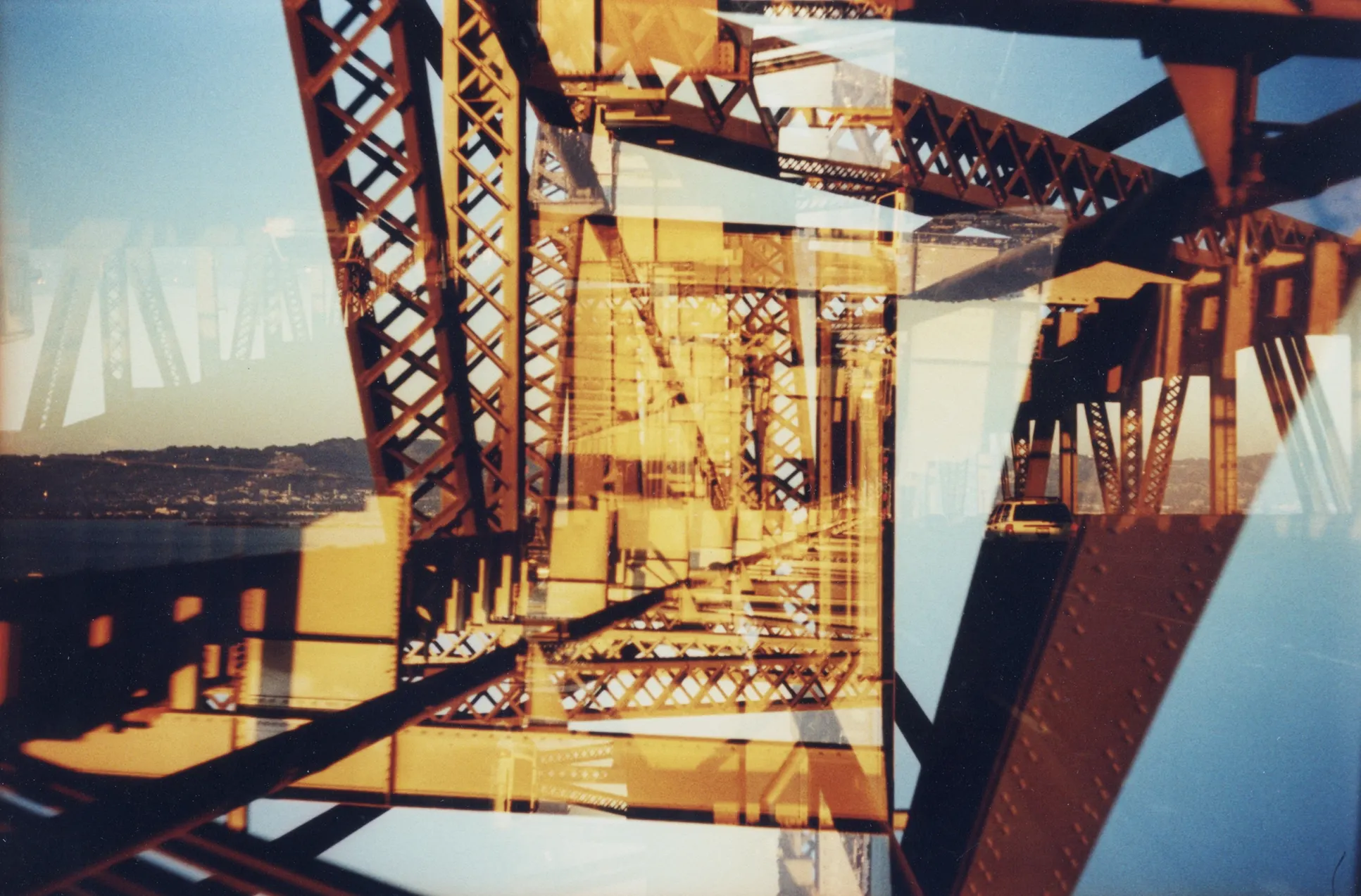 Multiple-exposure film photograph layering orange-red steel bridge trusses against blue sky and distant hills.