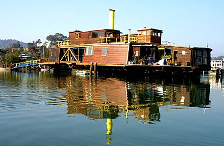 A large weathered wooden houseboat with a yellow smokestack floating on calm water, reflected below, with hills behind.