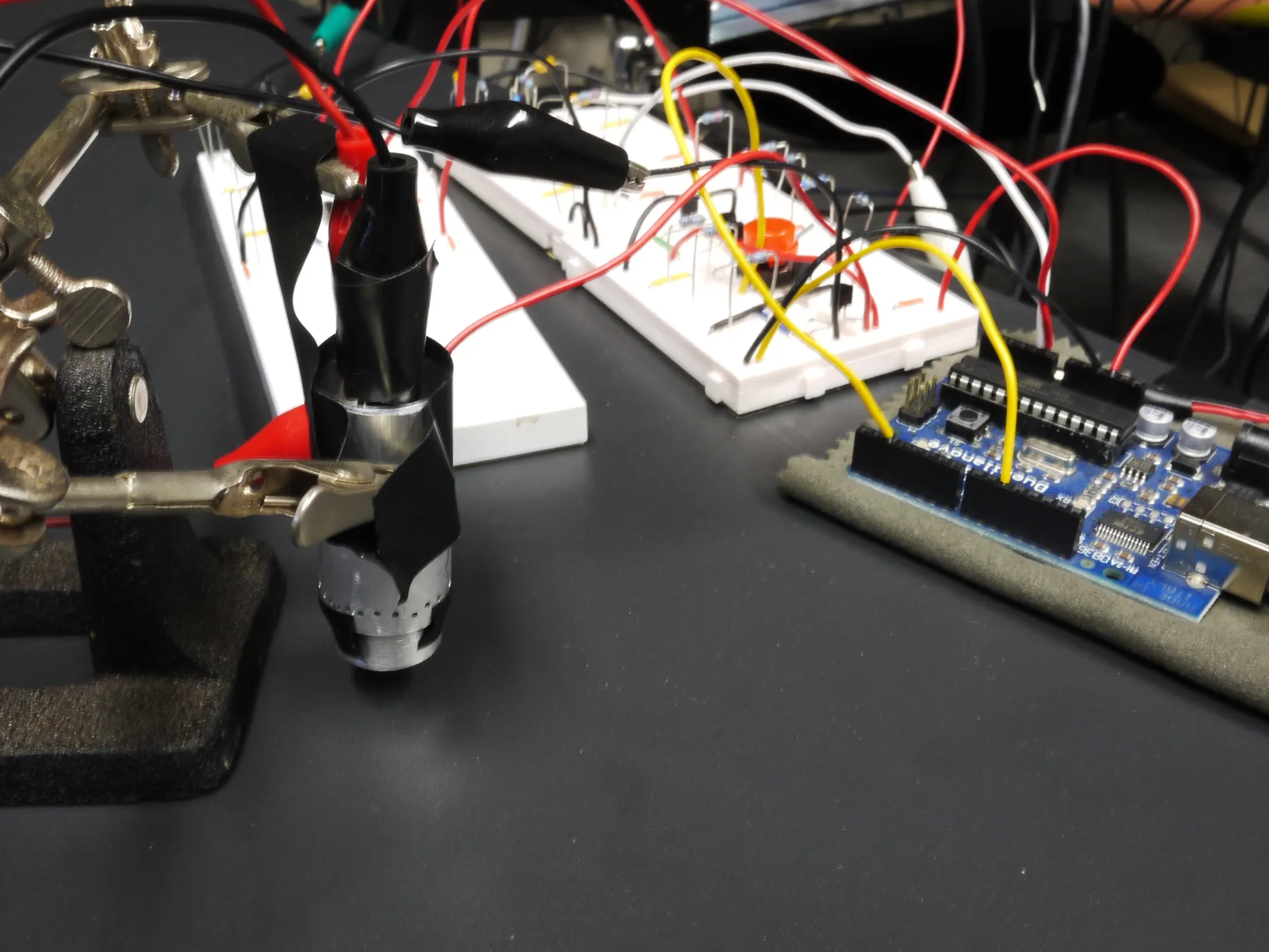 Electronics workbench with a helping-hands clamp holding a taped component, breadboards with red and yellow jumper wires, and a blue microcontroller board.