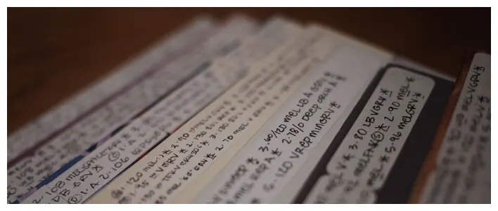Stacked handwritten index cards or paper strips fanned out on a dark surface, covered in small notations.