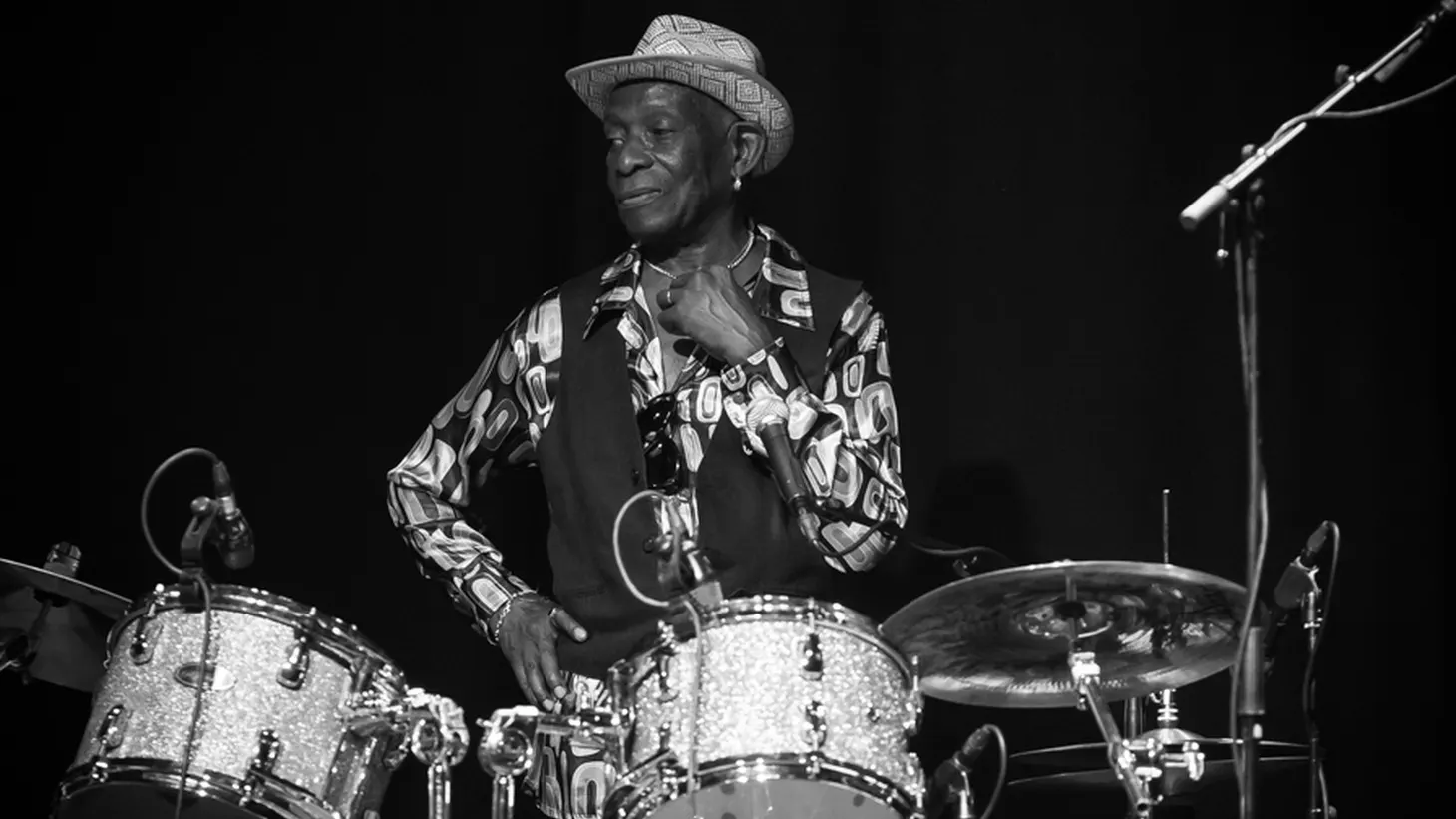 Black-and-white photo of a drummer in a straw hat and patterned shirt seated behind a sparkle-finish kit with cymbals and mics.